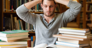 un homme est assis à une table dans une bibliothèque, entouré de nombreuses piles de livres. Il a les deux mains sur la tête, suggérant qu'il est stressé ou dépassé par la quantité de travail ou d'étude.