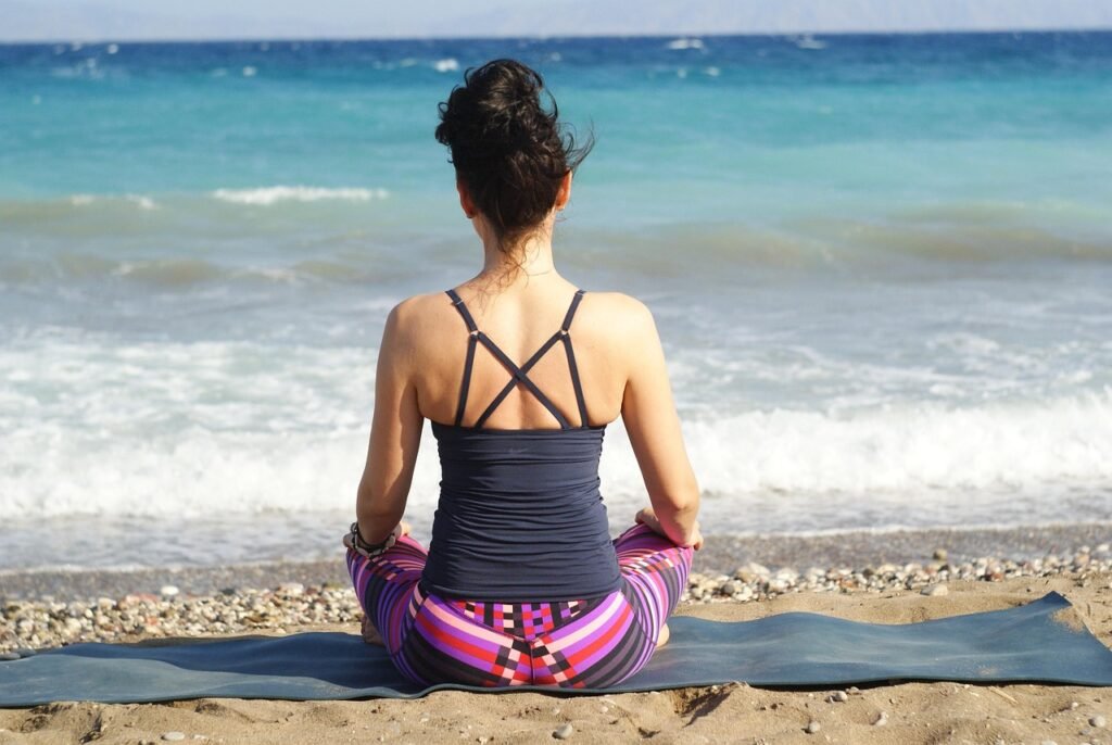 Une personne en méditation sur un tapis de yoga, assise face à la mer, avec des vagues douces en arrière-plan, portant des vêtements colorés, illustrant une méthode naturelle pour réduire le stress et améliorer la concentration avant les examens.