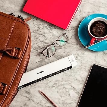 Un scanner portable Brother blanc sur un bureau avec sac, lunettes, tasse de café et tablette.