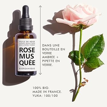 Bouteille d'huile de rose musquée bioorganique avec pipette en verre, placée à côté d'une rose rose pâle et d'une feuille verte. Étiquette indiquant "Huile de Rose Bio & Extra Vierge", "Dans Une Bouteille En Verre + Pipette En Verre", "100% Bio In France", et "YUKA: 100/100".