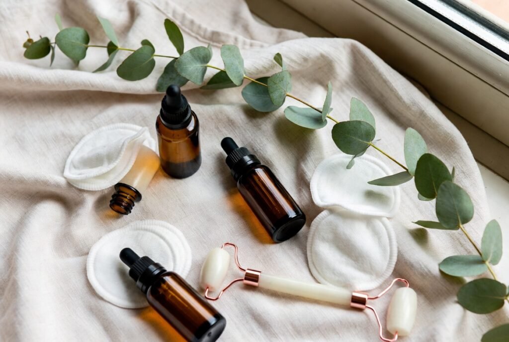 Composition de produits de soin pour la peau : flacons compte-gouttes en verre ambré, cotons démaquillants, roller facial en jade blanc avec poignées rose gold, et branches d'eucalyptus sur un tissu beige froissé près d'une fenêtre.