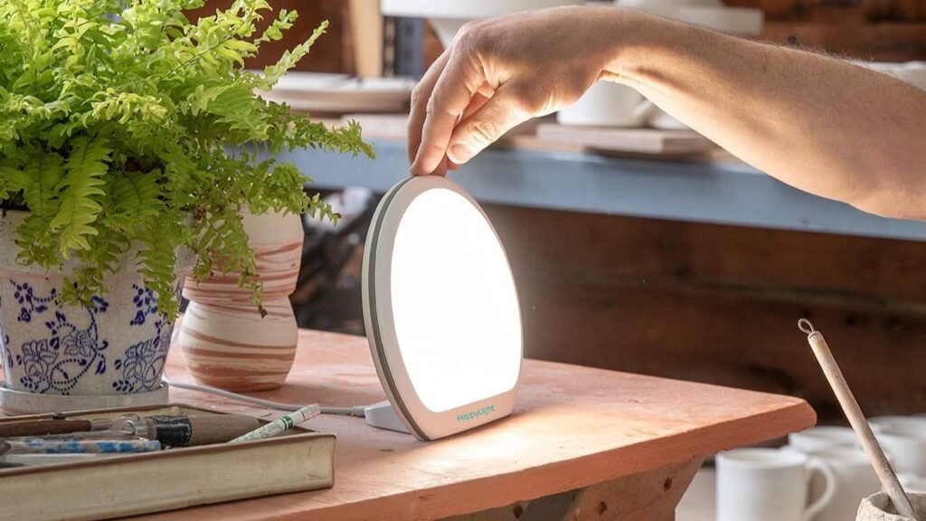 Lampe de luminothérapie Verilux HappyLight Alba allumée sur une table en bois, une main l’active à côté d’une plante verte