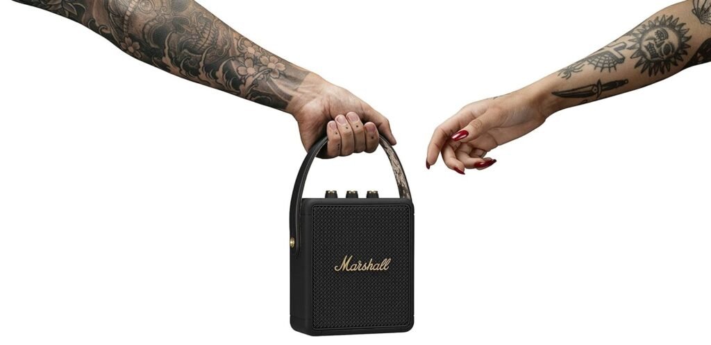 Enceinte portable Marshall : cadeaux tech couple, tenue par deux bras tatoués se passant l'objet sur fond blanc.